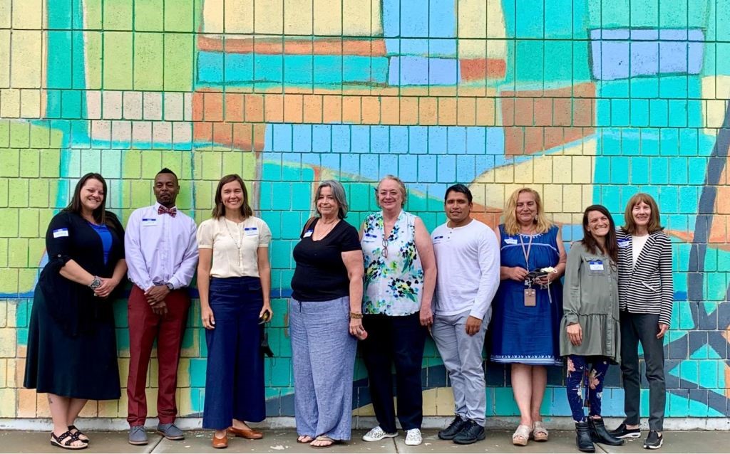 Seven members of the food equity resident advisors program stand in front of a colorful brick wall with Mayor Mendenhall and a project consultant.