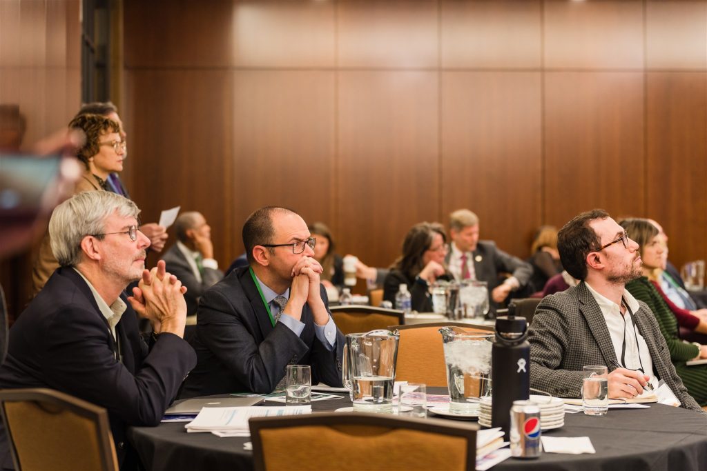 Conference attendees sit at a table listening to a presentation.