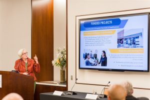 Former Mayor Hager-Smith presenting at a podium to an audience with visual powerpoint on the right.