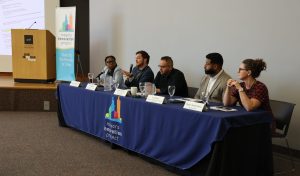 Five panelists sit at a speaker table. The panelist second from the left, Ian Fletcher, holds the microphone and is speaking to the audience.
