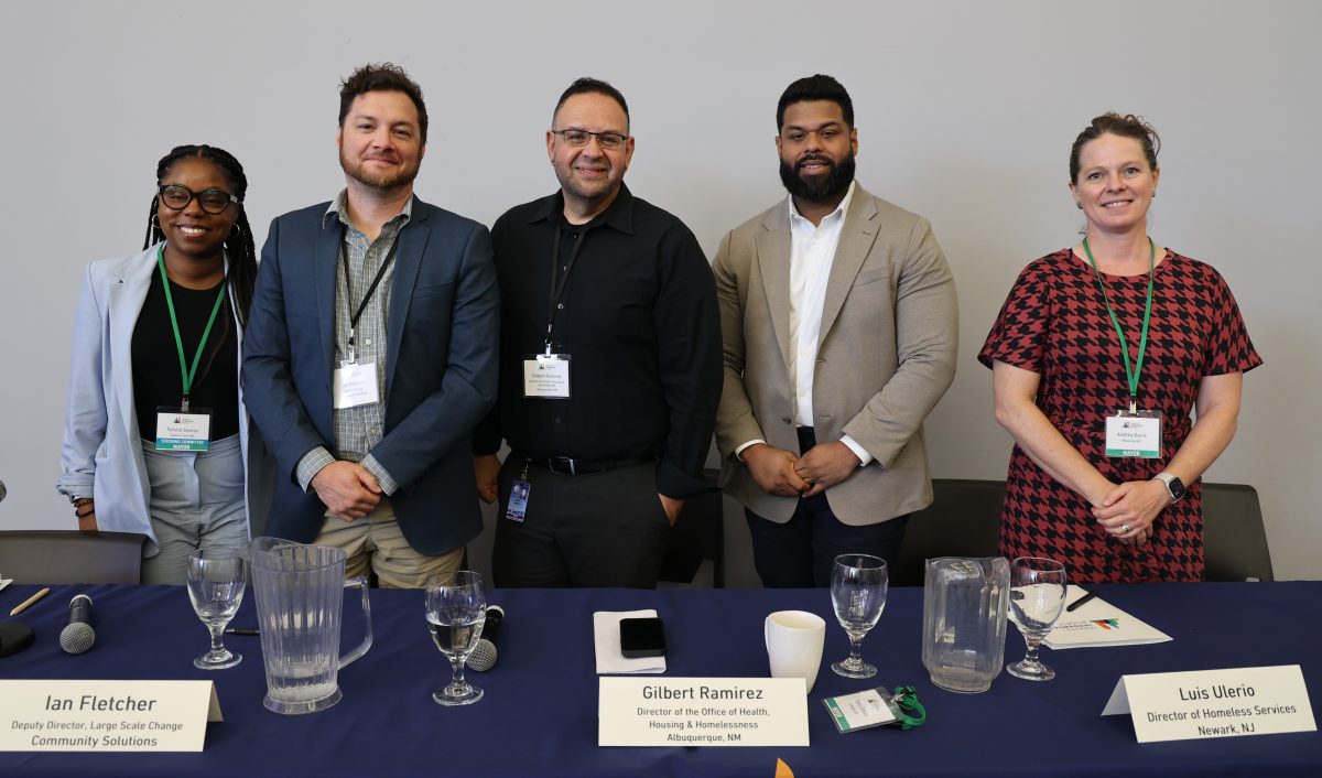 Five people stand behind a podium. From left to right: Panelists Mayor Talisha Searcy (Takoma Park, MD), Ian Fletcher (Community Solutions), Director Gilbert Ramirez (Albuquerque, NM), Director Luis Ulerio (Newark, NJ), and Mayor Angela Davis (Missoula, MT).