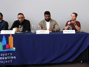 Three panelists sit at a speaker table. On the far right, Mayor Davis holds the microphone and speaks to the audience.