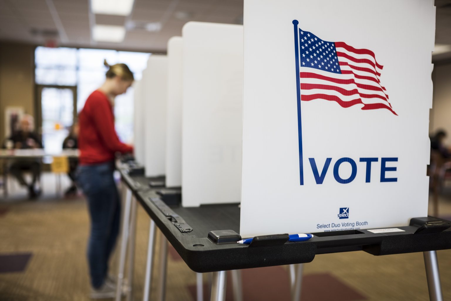 A voting booth is seen in focus, while voters are lined up out of focus